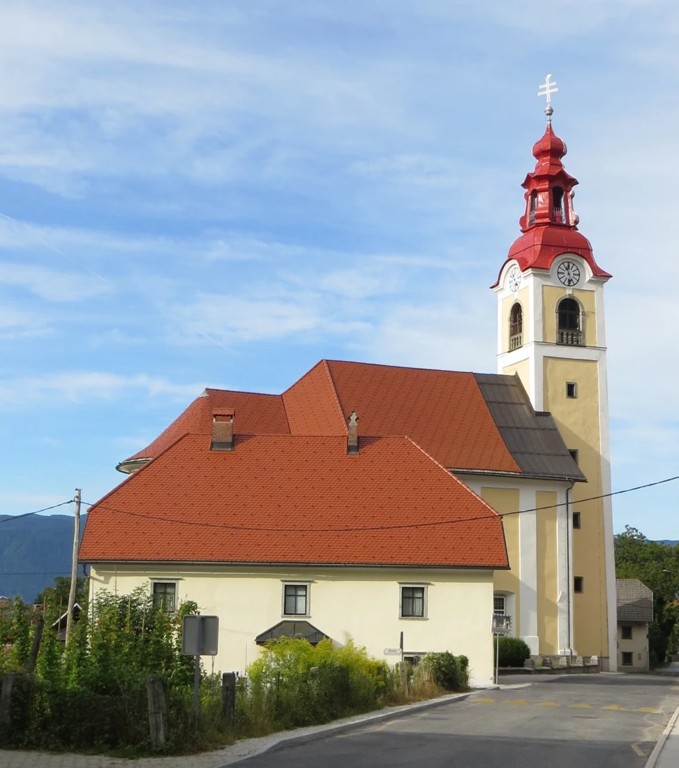 Svetla cerkev sv. Urha v Begunjah z rdečo streho in visokim zvonikom ob cesti.