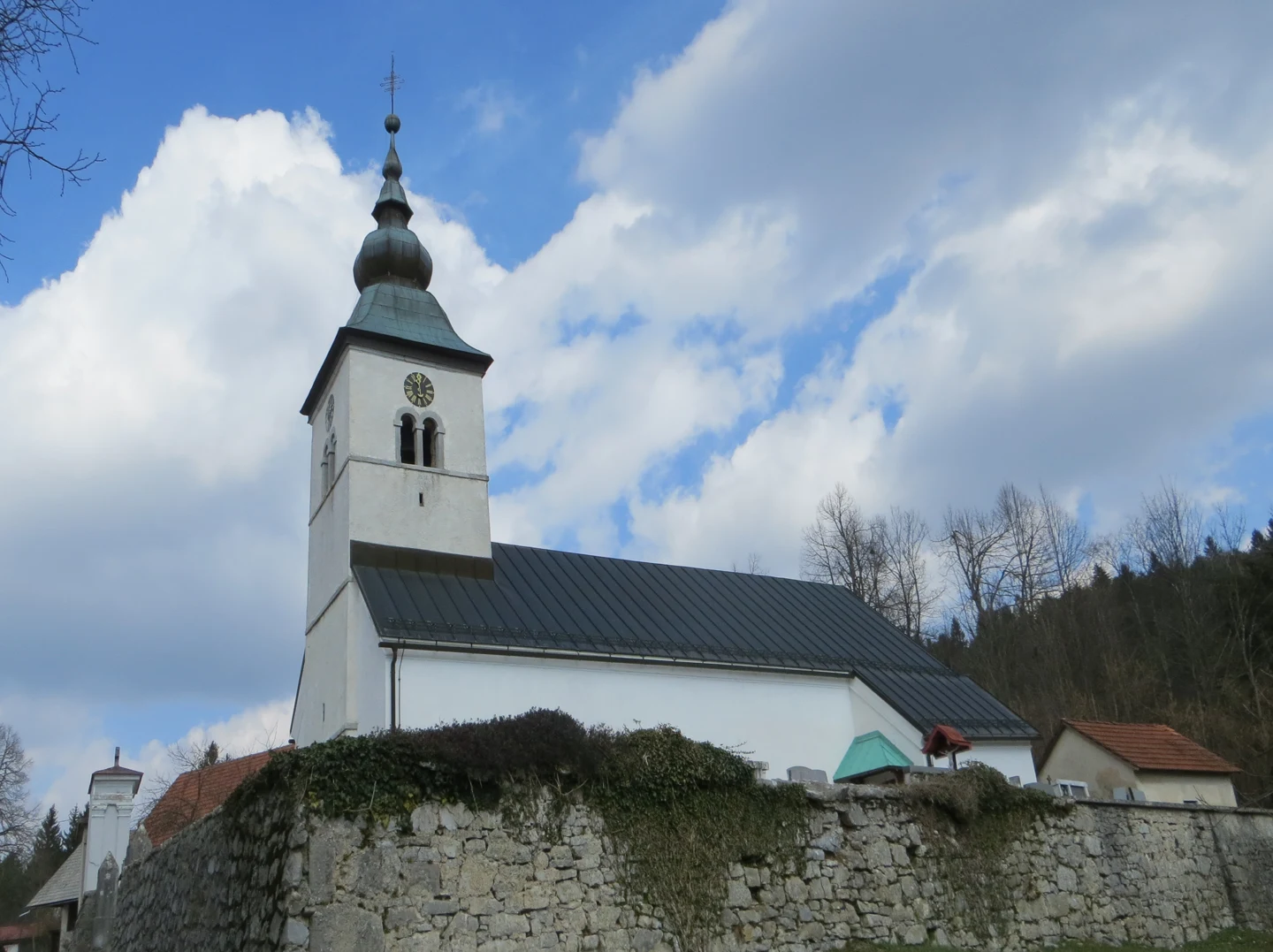 Cerkev Marijinega vnebovzetja v Bezuljaku nad pokopališčem, s kvadratnim zvonikom in čebulasto streho.
