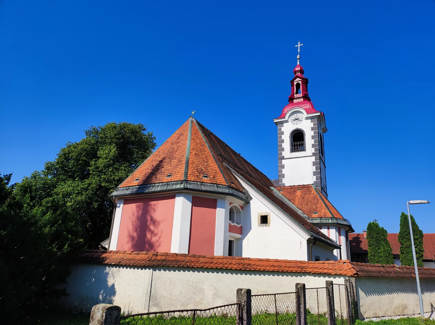 Cerkev sv. Mohorja in Fortunata v Grobljah z opečno streho, belo-rožnatimi stenami in zvonikom za obzidjem.