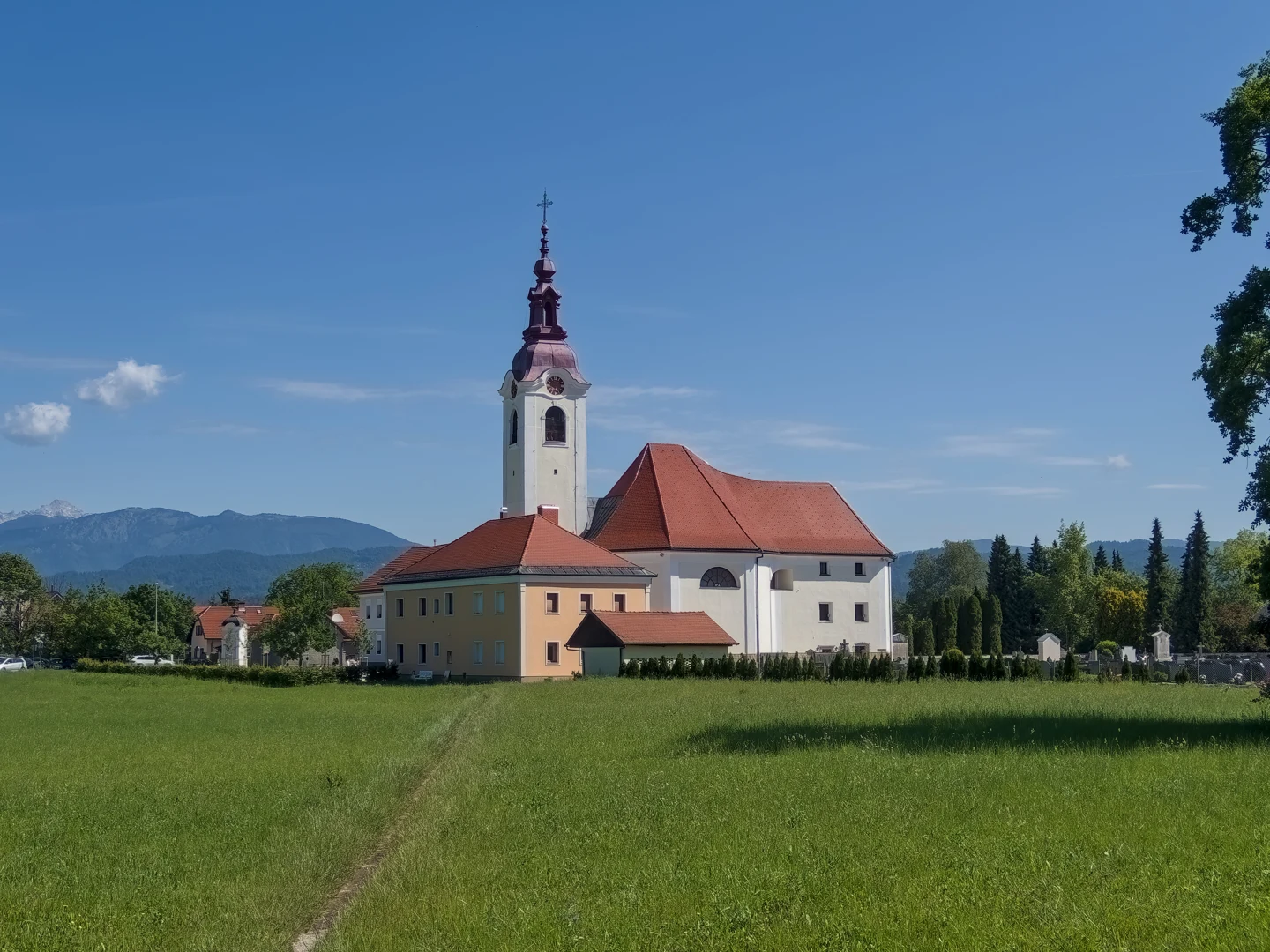 Cerkev sv. Martina v Dobu z visokim zvonikom in razgibano opečno streho, ob njej nižja stavba in pokopališče; v ospredju travnik.