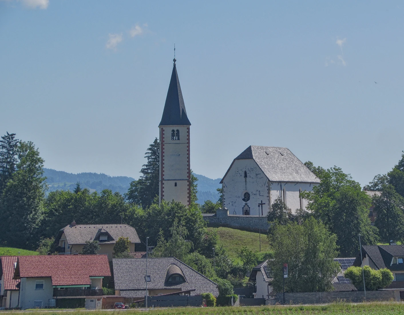 Cerkev na Krtini s samostojnim visokim zvonikom, svetlim pročeljem in kamnitim obzidjem na griču nad hišami.