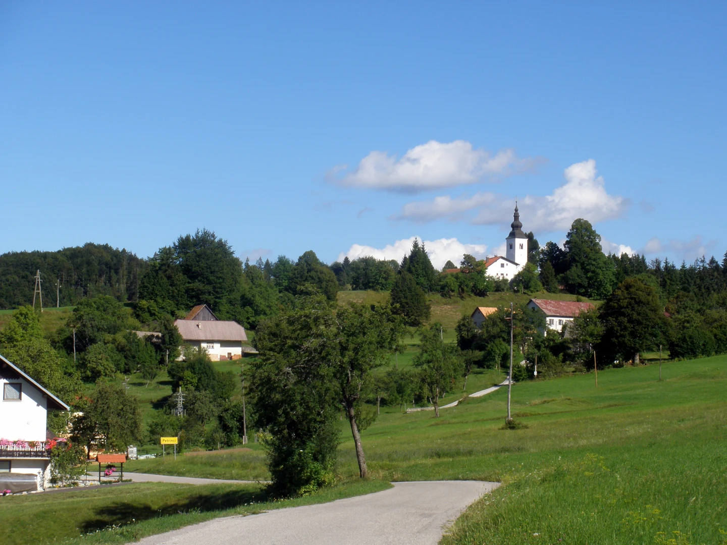 Bela cerkev Marije Snežne z baročnim zvonikom nad zelenim pobočjem in hišami Petrincev, v ospredju tabla Petrinci.