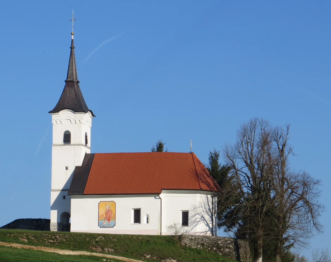 Bočni pogled na cerkev sv. Janeza Evangelista v Gorenji vasi z zunanjim poslikanim svetnikom in zvonikom, april 2018