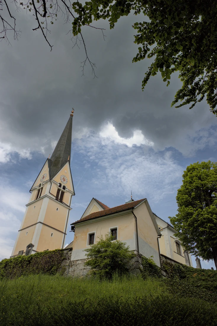 Cerkev Marijinega rojstva na Homcu in ločen zvonik s strmo koničasto streho, nad travnatim pobočjem pod oblačnim nebom.