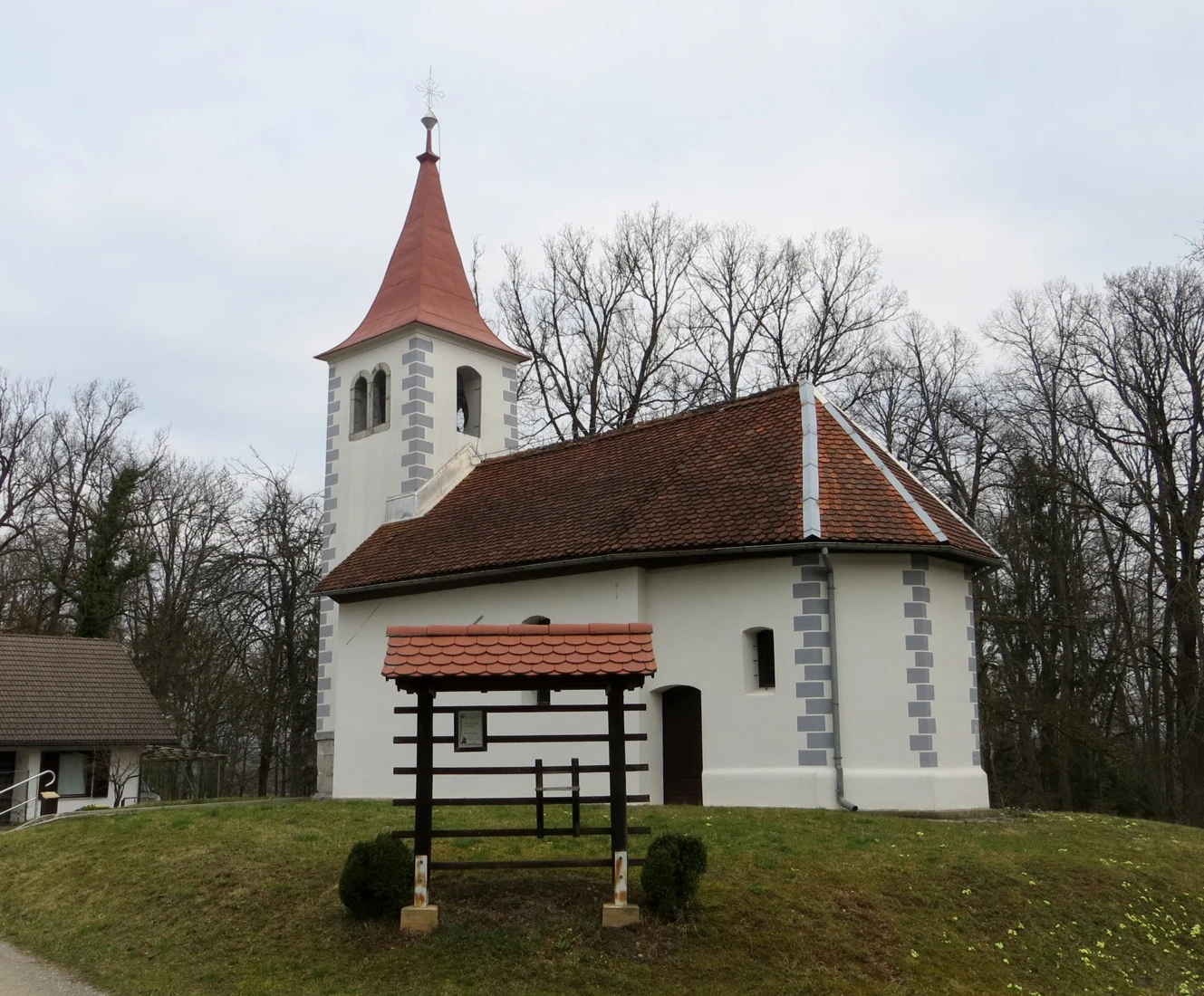 Svetla cerkev sv. Urha z rdeče kritim zvonikom in prezbiterijem na Kremenici; pred njo leseno znamenje.