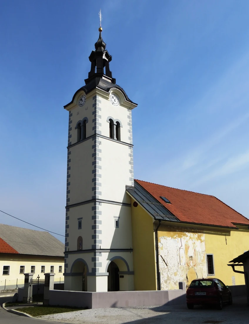 Zvonik z uro in temno baročno streho ob rumeni ladji cerkve v Mateni, obodni zid in vhod.