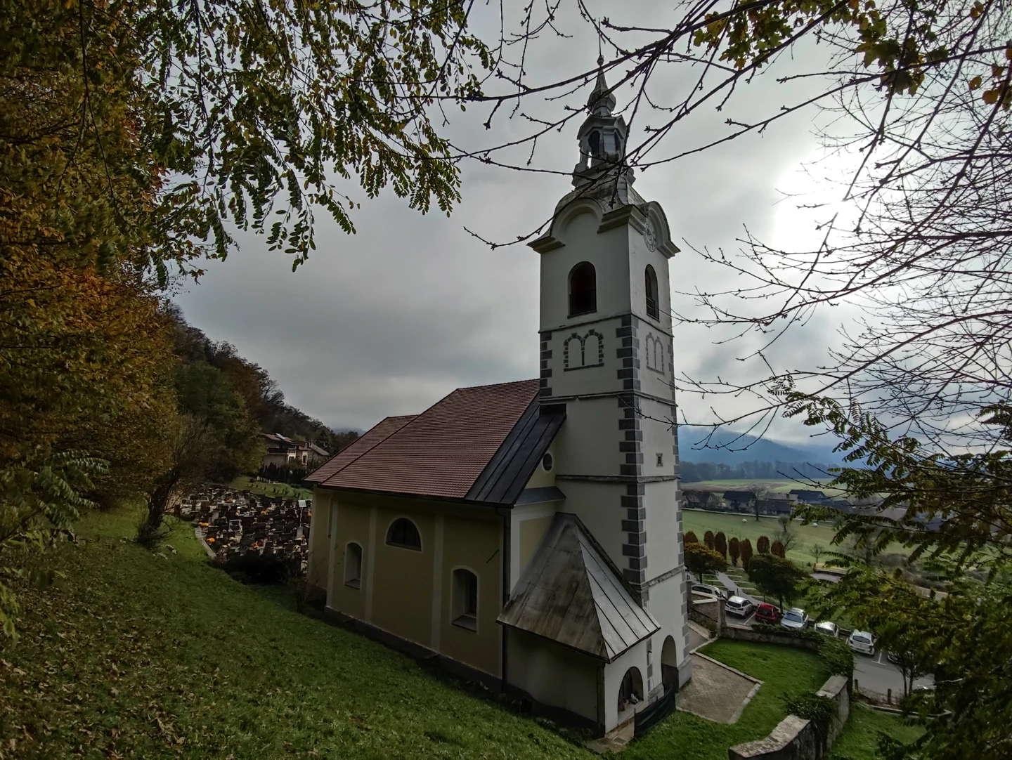 Cerkev sv. Jurija v Ihanu z visokim zvonikom in opečno streho, med jesenskim drevjem; ob strani pokopališče, v ozadju polja.