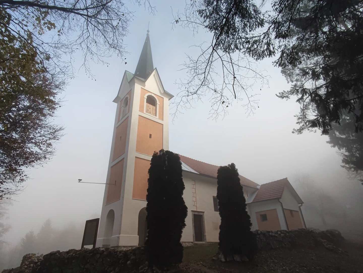 Cerkev sv. Nikolaja na Goropečah z visokim zvonikom, svetlimi stenami in rdečo streho med drevesi v megli.