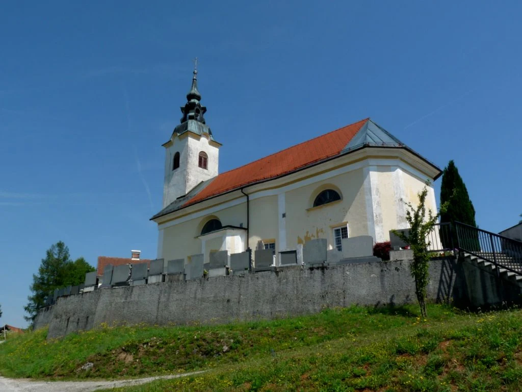 Svetla cerkev na Kopanju z rdečo streho in zvonikom nad pokopališkim zidom ter vrsto nagrobnikov.