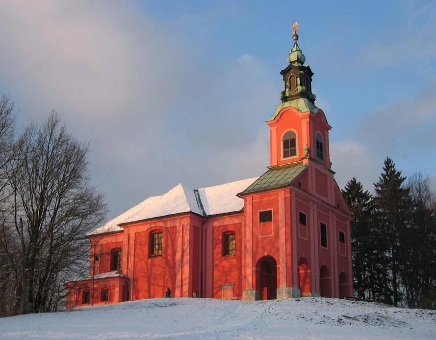 Rdeča cerkev Marijinega obiskanja na Rožniku z zvonikom, zasneženo streho in drevjem.