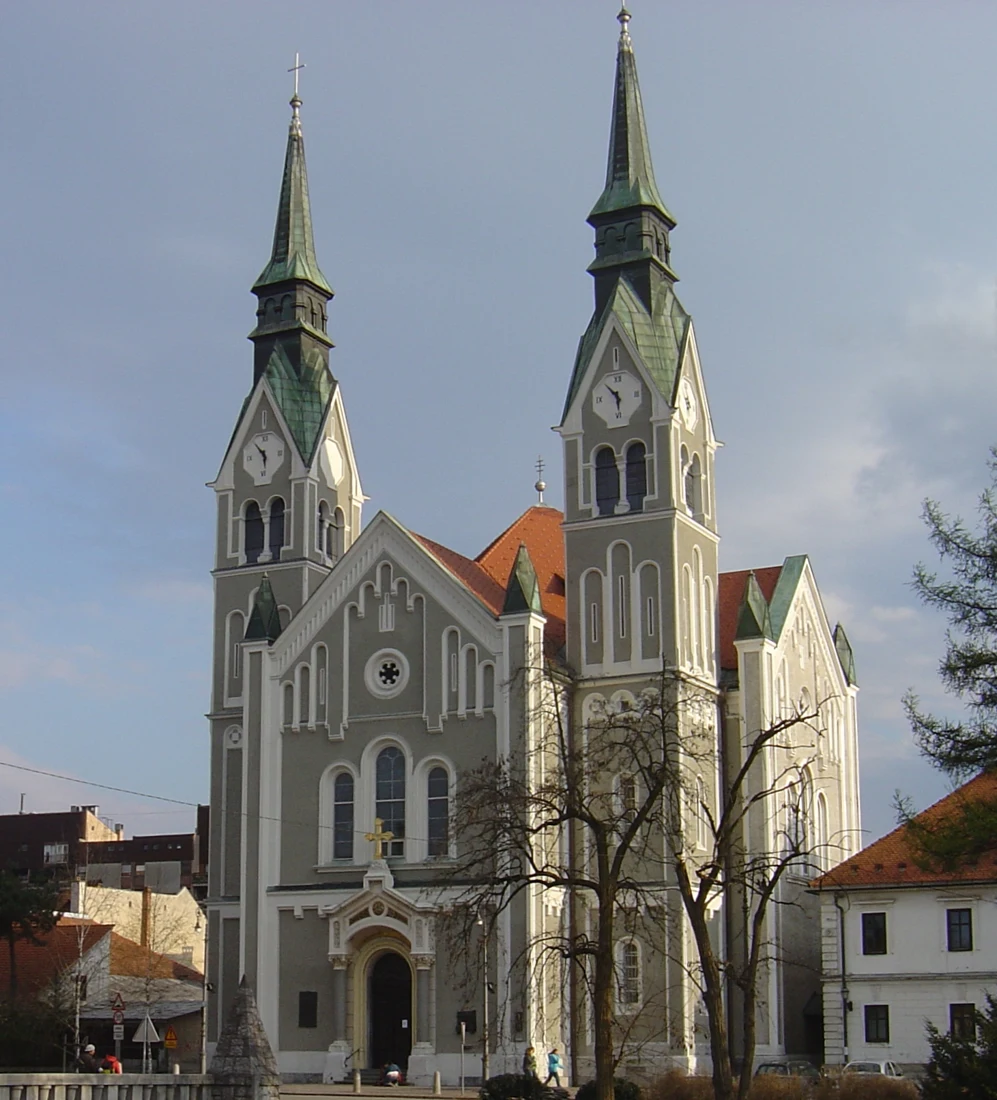 Pročelje trnovske cerkve sv. Janeza Krstnika z visokima zvonikoma, urama in drevjem pred vhodom.