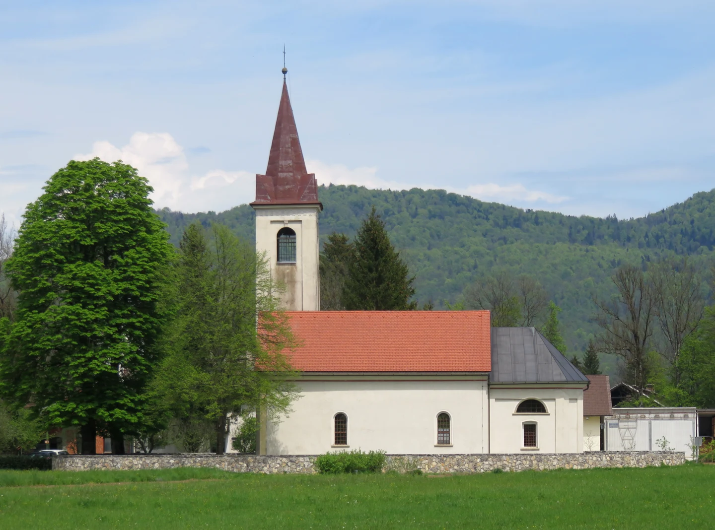Bočna stran cerkve Imena Marijinega v Goriči vasi z rdečo streho, visokim zvonikom in nižjim prezbiterijem za kamnitim zidom.