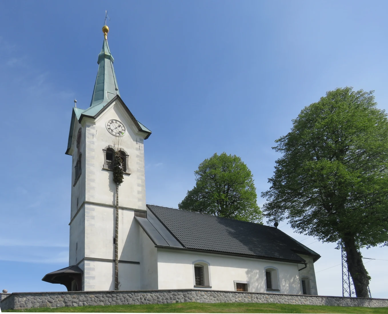 Svetla cerkev sv. Hieronima na Petkovcu z visokim zvonikom z uro, ladjo in nizkim kamnitim obzidjem.