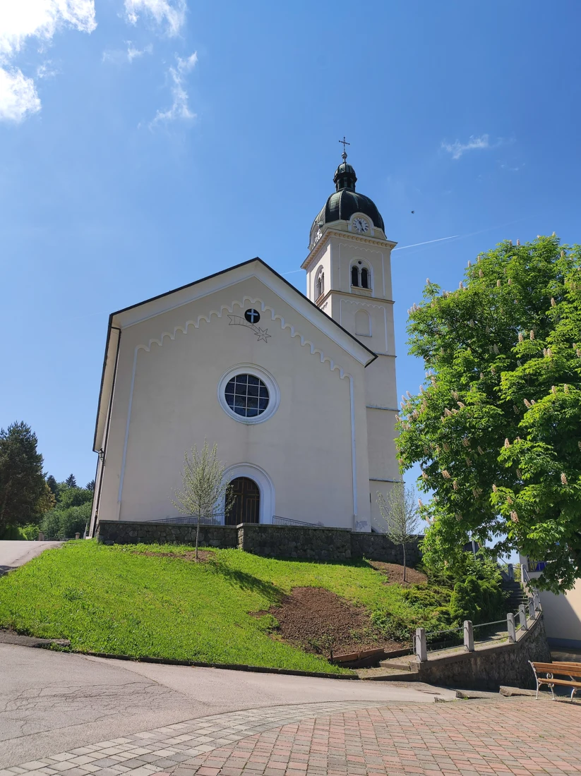 Severno pročelje cerkve sv. Urha v Smledniku z rozeto in zvonikom nad travnato brežino.