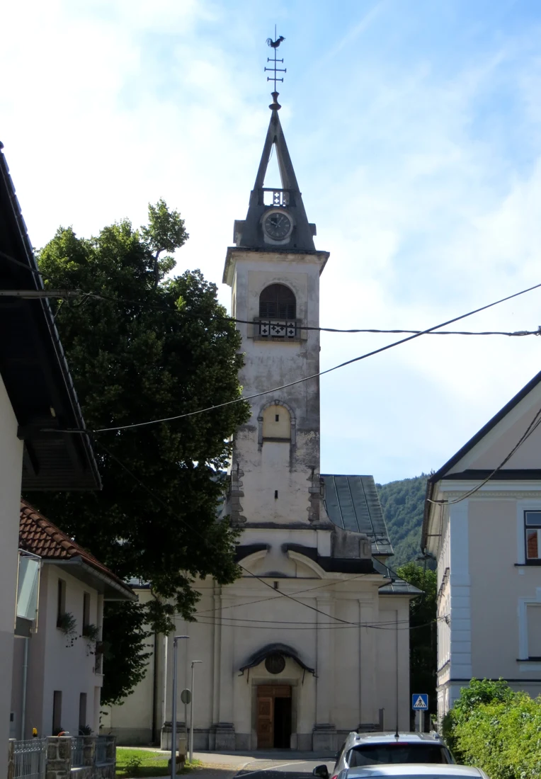 Pročelje cerkve sv. Marije Magdalene v Sodražici z visokim zvonikom, uro in petelinom, med sosednjima stavbama.