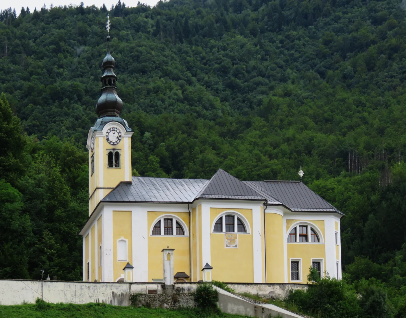 Rumeno-bela cerkev sv. Martina z zvonikom in čebulasto streho pod gozdnatim pobočjem.