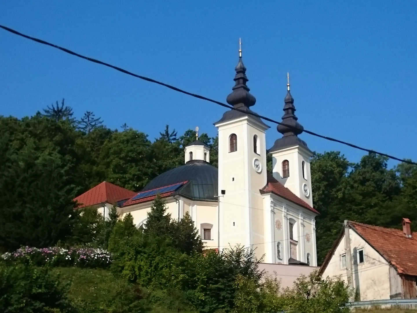 Svetla cerkev v Tomišlju z dvema zvonikoma z urama in temno kupolo na gozdnatem pobočju.