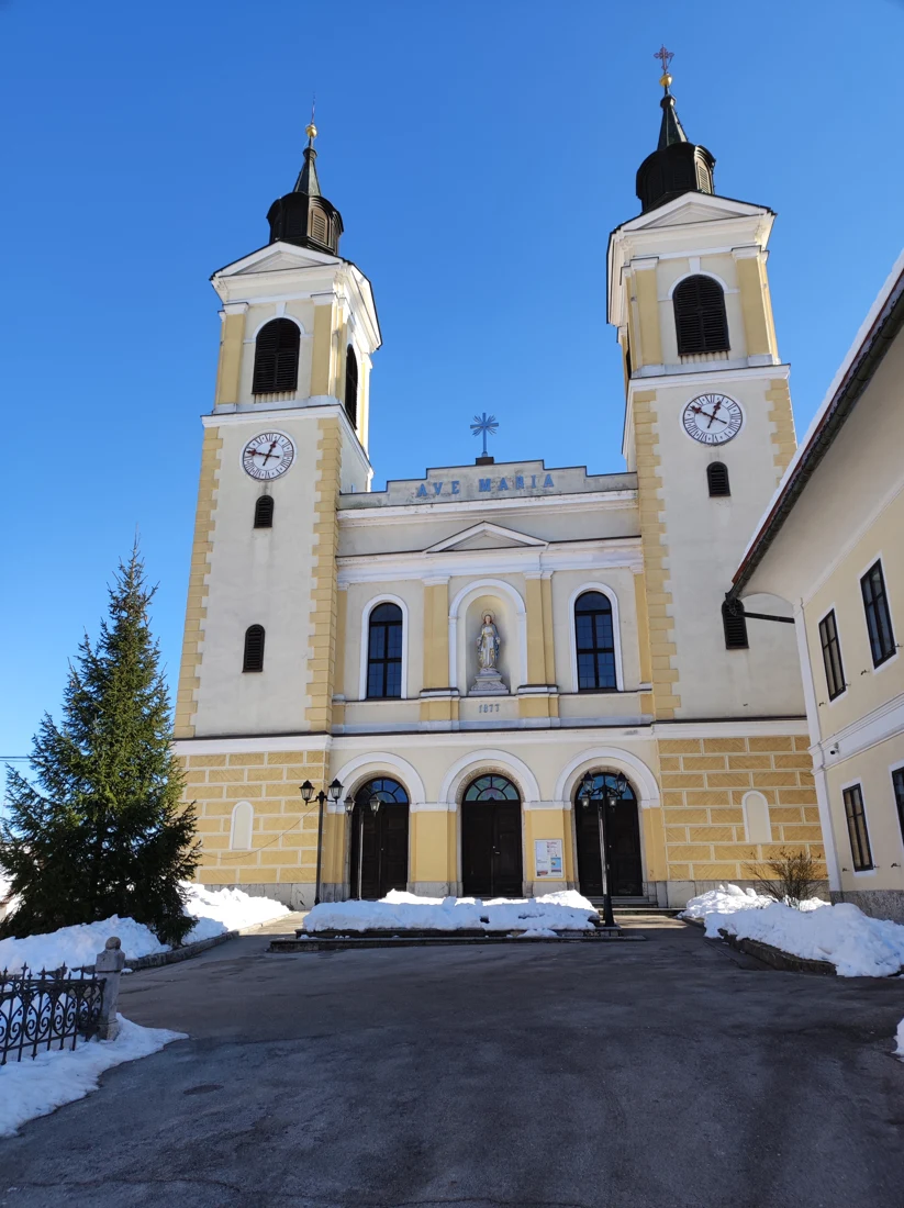 Pročelje cerkve Marijinega rojstva v Velikih Laščah z dvema zvonikoma, urama, napisom AVE MARIA in tremi vhodi ob zasneženih stopnicah.