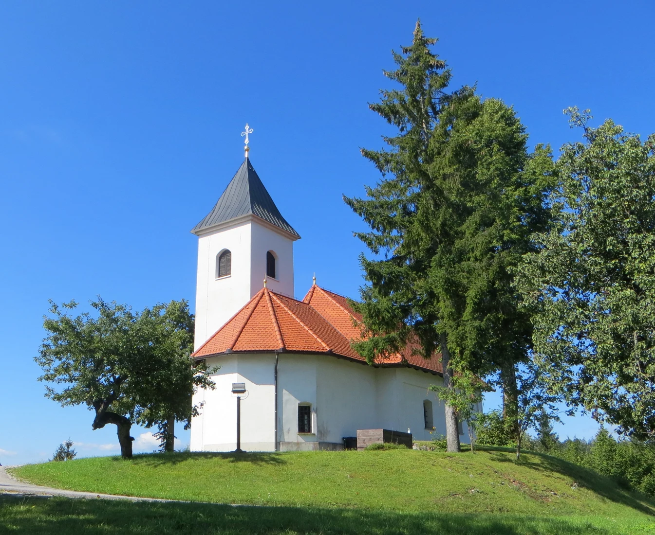Bela cerkev sv. Ožbolta na Lužarjih z visokim zvonikom, rdečo streho in drevjem na travnati vzpetini.