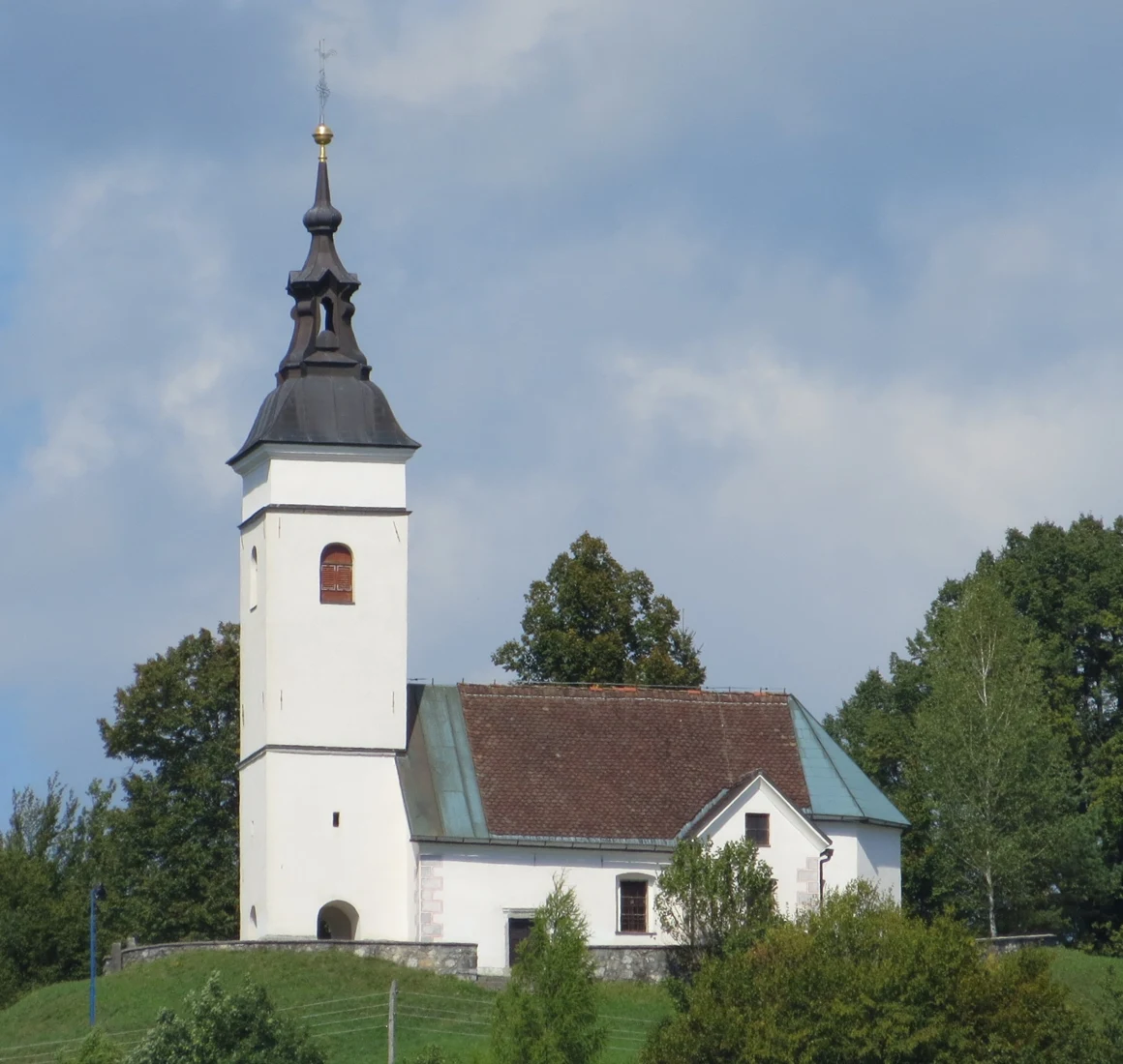 Bela cerkev sv. Lenarta v Mali Ligojni z visokim zvonikom in nizkim obzidjem na travnatem griču.