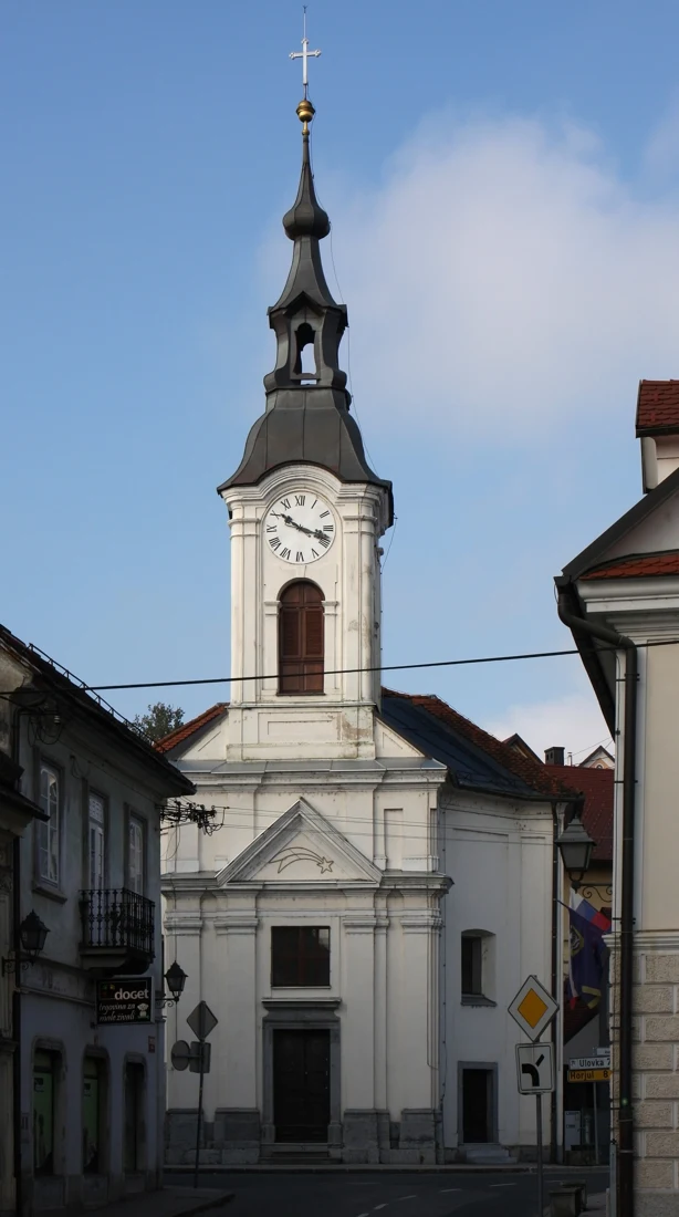 Svetlo pročelje cerkve sv. Lenarta med mestnima hišama, z osrednjim zvonikom, uro, kamnitim vhodom in stopnicami.