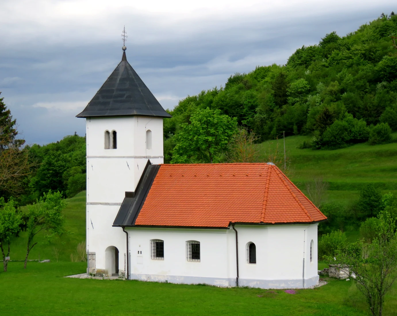 Bela cerkev sv. Lenarta z rdečo streho in zvonikom ob zelenem travniku ter gozdu na Gornjem Igu.