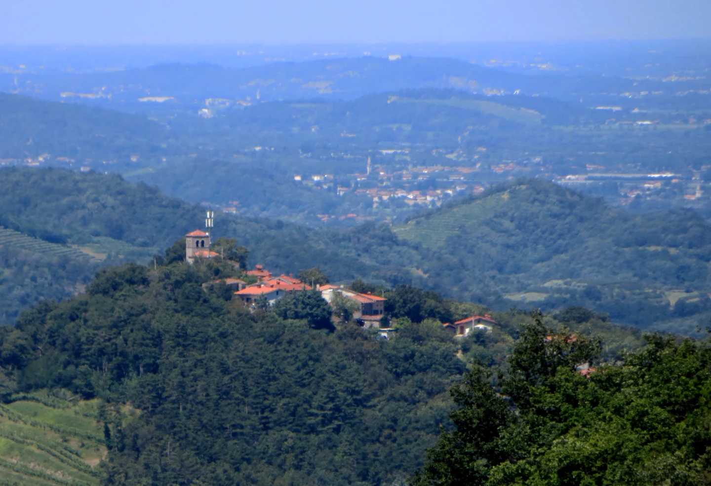 Pogled na Sveti Martin nad Brjami s cerkvenim zvonikom na levem grebenu in hišami med zelenjem.