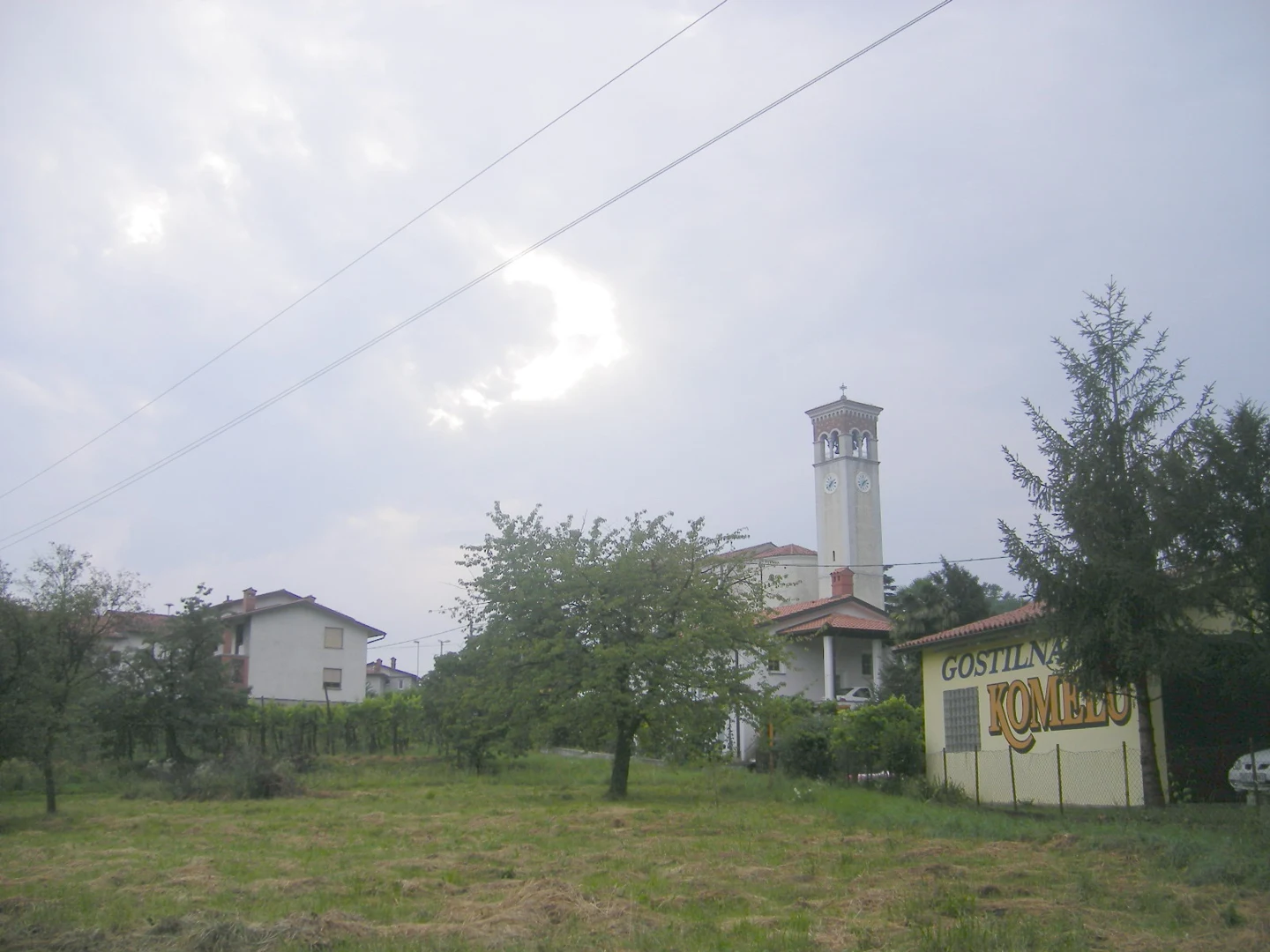 Zvonik cerkve Marije Tolažnice v Kromberku in del cerkvene stavbe za drevjem ob travniku.