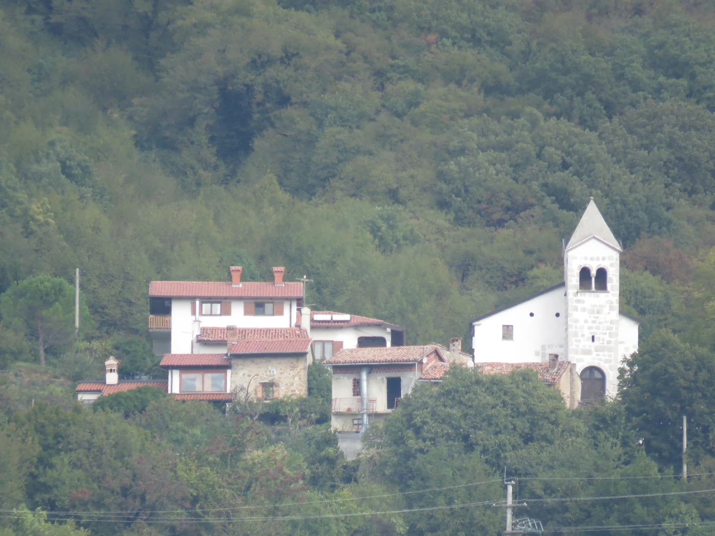 Cerkev sv. Mihaela s kamnitim zvonikom ob vaških hišah pod gozdnatim pobočjem.